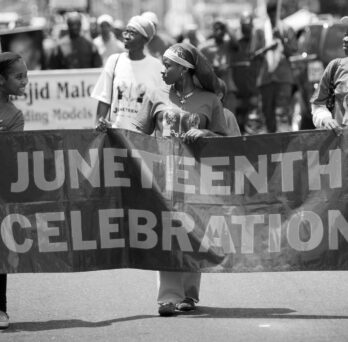 Black-and-white photo of people marching in a Juneteenth celebration while holding a large banner that reads “Juneteenth Celebration.” 