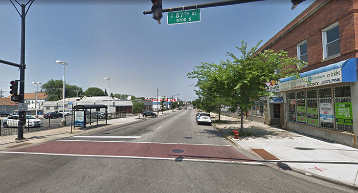 Empty intersection with a storefront and one tree on the right, and a parking lot and bus stop on the left.