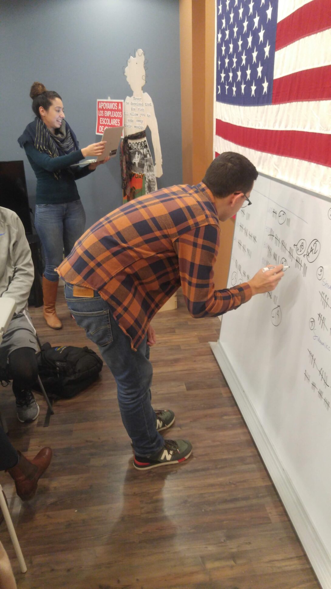 Man writing on white board while woman reads off of laptop. American flag in background.