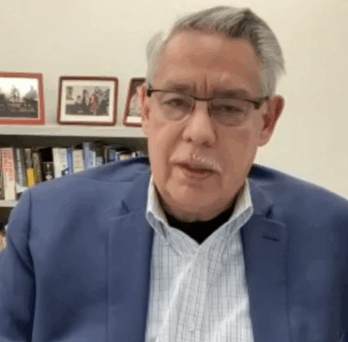 Man speaking on camera in office setting, seated before bookshelf and filing cabinets, wearing glasses and blue blazer. 