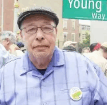 José ‘Cha Cha’ Jimenez, an older man wearing glasses, a gray flat cap, and a light blue shirt, stands at an outdoor community gathering. Behind him is a green street sign reading ‘Young Lords Way,’ with several people and a brick building visible in the background. 