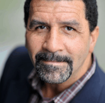 Portrait of Dr. Stephen Small, a man with short curly hair and a salt-and-pepper goatee, wearing a blazer and patterned shirt, looking toward the camera. 