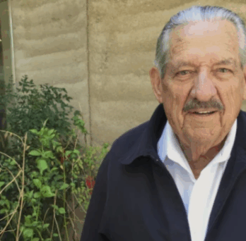 Portrait of Senator Fred Harris, an elderly man with gray hair and a mustache, standing outdoors near a concrete wall and greenery. 