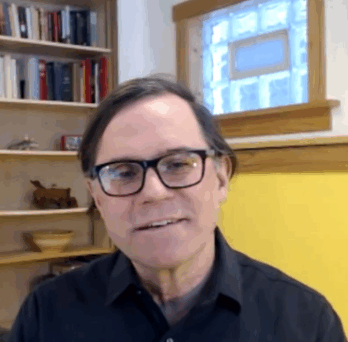 Man speaking on camera in home office setting, with bookshelves, window, and decorative items visible in background. 