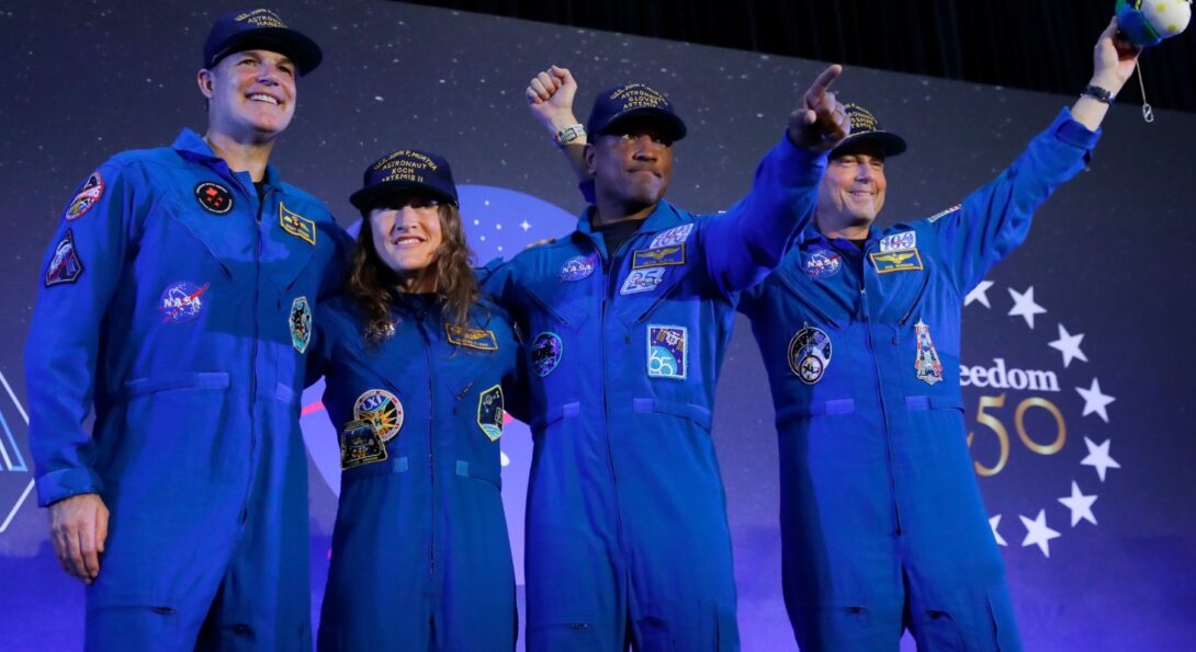 Four individuals in blue flight suits and caps stand on stage, smiling and raising their hands, with a backdrop featuring stars and “Freedom 50.”