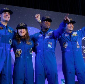 Four individuals in blue flight suits and caps stand on stage, smiling and raising their hands, with a backdrop featuring stars and “Freedom 50.” 