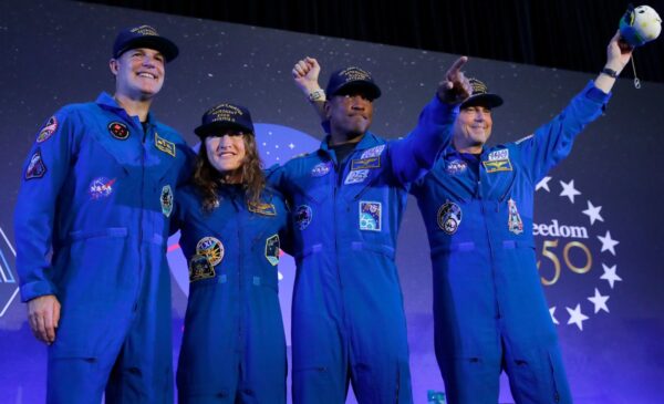 Four individuals in blue flight suits and caps stand on stage, smiling and raising their hands, with a backdrop featuring stars and “Freedom 50.”