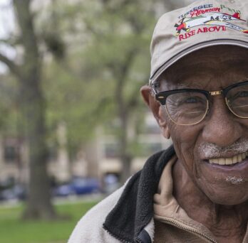 Portrait of civil rights activist, educator, and historian Timuel Black smiling outdoors while wearing glasses and a baseball cap. 
