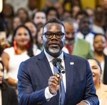 Chicago Mayor Brandon Johnson speaks before a large crowd during a public event, standing at a podium with supporters behind him. 