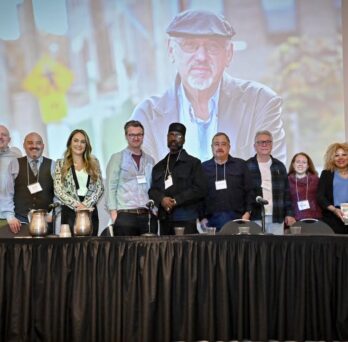 Panelists and speakers stand together onstage during an event honoring John Hagedorn, with his portrait displayed on a large screen behind them. 