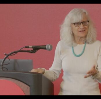 Speaker Jane Caputi stands at a podium during a Great Cities Institute event, presenting on ecofeminism, the Anthropocene, and environmental justice. 
