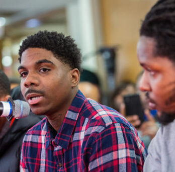 Young man speaking into a microphone at a public hearing, seated beside another attendee. 