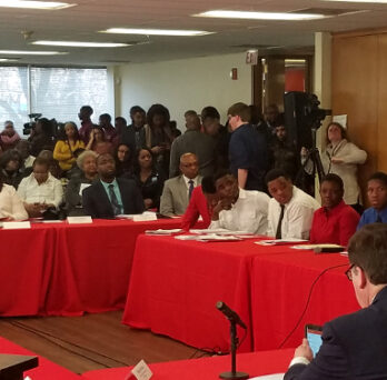 Audience members seated at red-covered tables during a public hearing or forum, with speakers and attendees gathered in the room. 