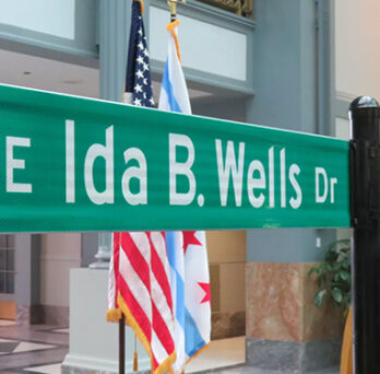 Ida B. Wells Drive street sign displayed indoors beside an American flag and podium. 