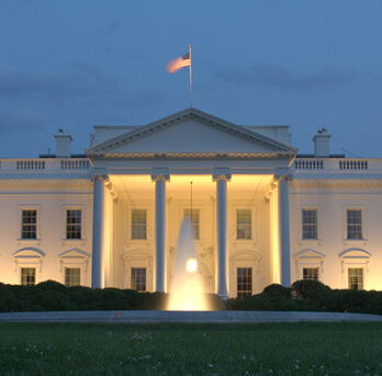 The White House illuminated at dusk, with columns lit and an American flag flying above the building. 