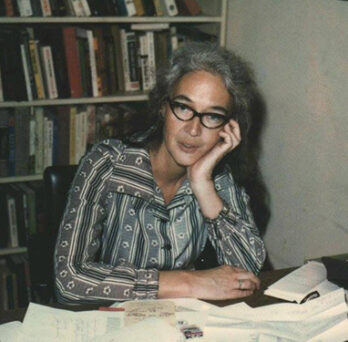 Older woman wearing glasses seated at a desk with papers, books, and shelves in the background. 