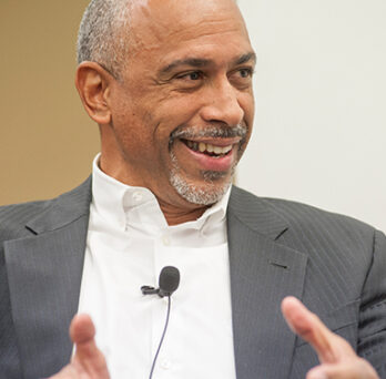 Pedro Noguera speaking at a lecture or panel, wearing a suit jacket and gesturing while seated near a microphone. 