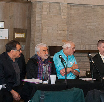Panelists seated at a table during a discussion on Latino gang structures, with a moderator standing at a podium. 