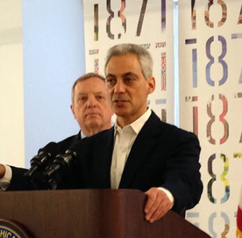Man speaking at a podium during an official announcement, with another man standing behind him and an American flag nearby. 