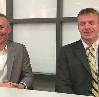 Two men in suits seated at a table in front of frosted glass windows. 