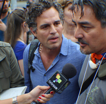 Man speaking into a microphone during a crowded outdoor interview, with reporters and attendees gathered around him. 