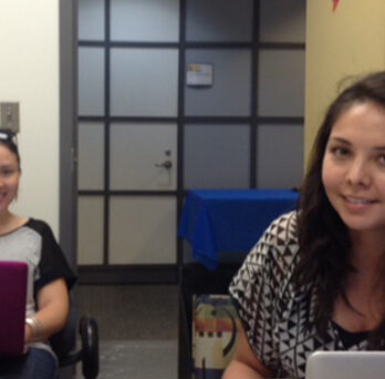 Two women working at laptops in an office or conference room, seated at tables with glass-paneled doors in the background. 