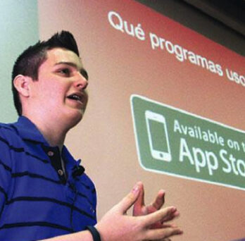 Teen presenter speaking in front of a projected slide showing an App Store graphic, during a talk about apps or youth entrepreneurship. 