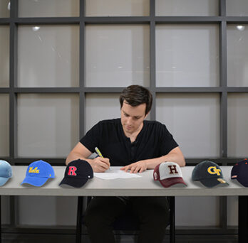 Person seated at a table signing papers, with university baseball caps lined up across the table. 