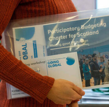 Person holding a printed Participatory Budgeting Chapter for Schools toolkit booklet in front of a window. 