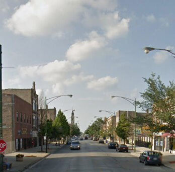 Street view of South Commercial Avenue with brick storefronts, parked cars, sidewalks, and neighborhood businesses under a cloudy sky. 