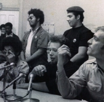 Black-and-white photo of activists seated and standing at a press conference, speaking into microphones. 