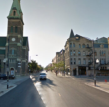 Street view of Pilsen with brick buildings, a church tower, and cars along a wide neighborhood road. 