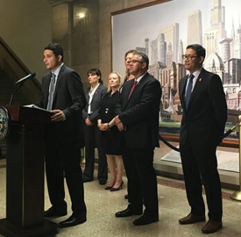 Speaker at a podium during a press conference, with several people standing behind him in front of a city mural. 