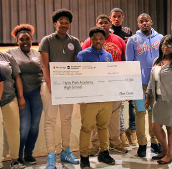 Group of students and adults standing on a stage, holding an oversized check for a school participatory budgeting project. 