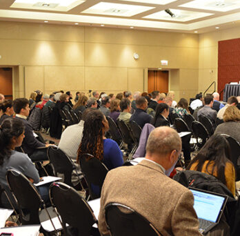 Large audience seated in a conference room facing a panel of speakers at the front of the room. 