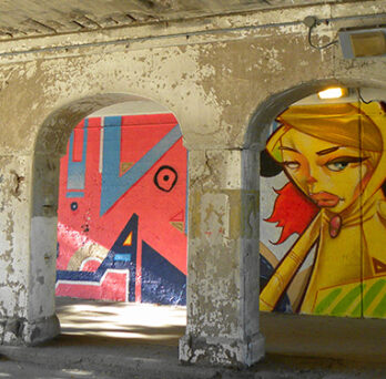Murals painted on the walls of a concrete viaduct, featuring colorful portrait-style artwork and abstract designs under arched openings. 