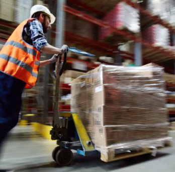 Warehouse worker wearing a hard hat and reflective vest pushes a pallet jack loaded with wrapped goods through a warehouse aisle. 