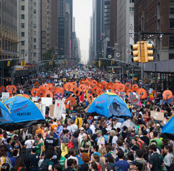 Large crowd marching through a city street during a climate demonstration, with banners, umbrellas, and tall buildings surrounding the march. 
