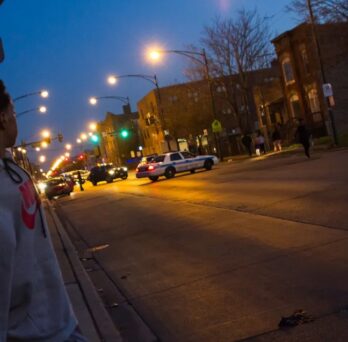 Person standing on a city sidewalk at night, looking toward police cars and streetlights along the road. 