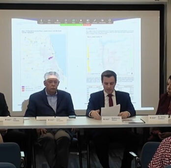Panel discussion with four speakers seated at a table in front of a projected map, with audience members seated in the foreground.